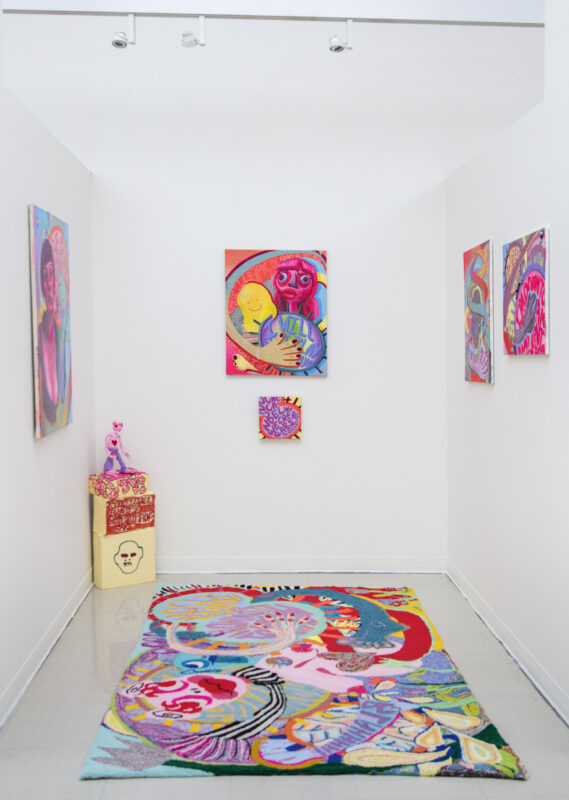 Installation including a colorful tufted rug, painted pedestal, a brightly painted clay figure and brightly colored paintings