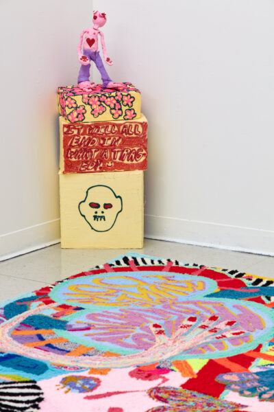 Installation including a colorful tufted rug, painted pedestal and a brightly painted clay figure