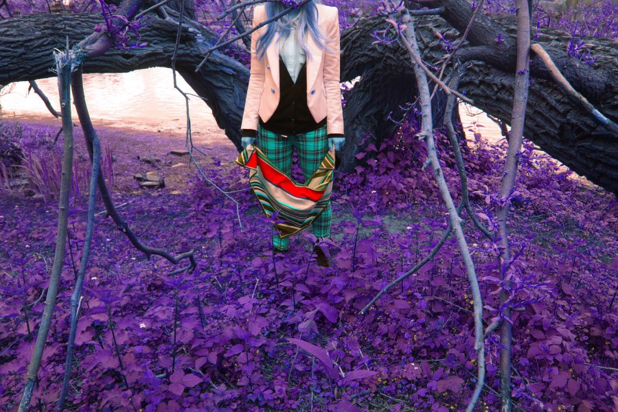 A girl in front of a tree and in the middle of tall grass. The photograph's colors are inversed as the grass is purple, the skin is blue, and all the other colors are inverted.