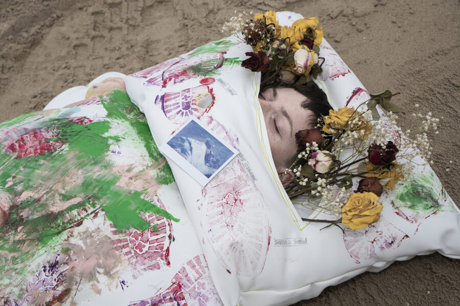 A woman with an abstract bed-like clothes standing in the middle of the photo by the beach, on the bed are paintings of colorful disorganized footprints.