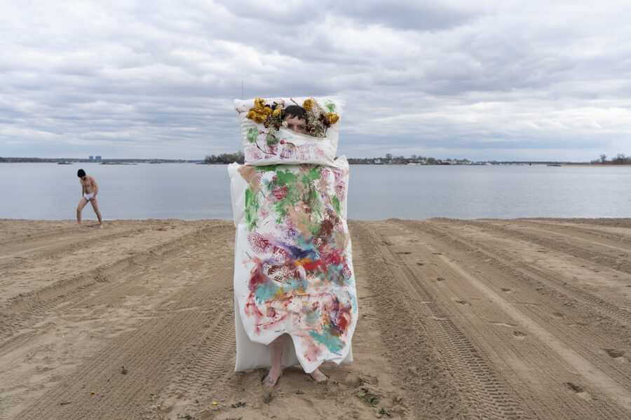 A woman with an abstract bed-like clothes standing in the middle of the photo by the beach, on the bed are paintings of colorful disorganized footprints.