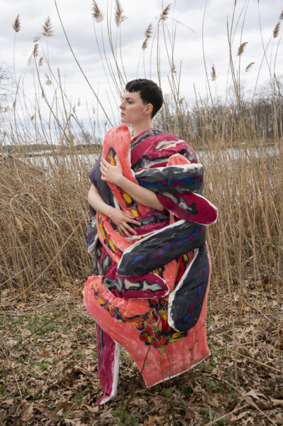 A skeleton hand made by fabrics crossing on the body of a woman. The fabric is a large wrapped piece with red, brown, and white colors.. The model stands looking to the left. In the background is a field with water in the distance.