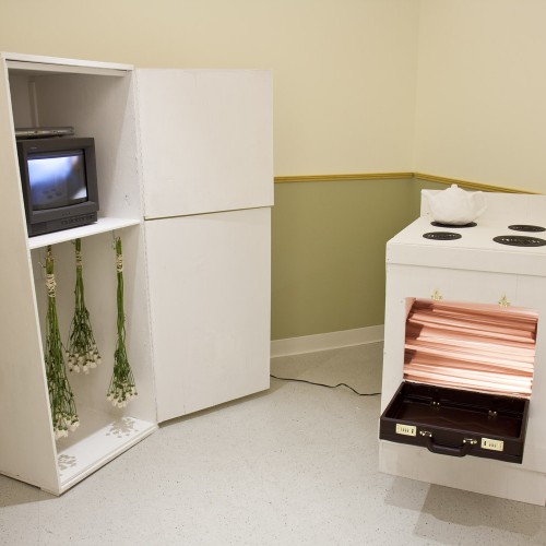 Installation view of a stove with a white tee-pot on it and the oven door open, a cabinet with two storage parts door open and on the top part is a TV with an image on it, on the bottom part are a few flower bouquet hung upside down