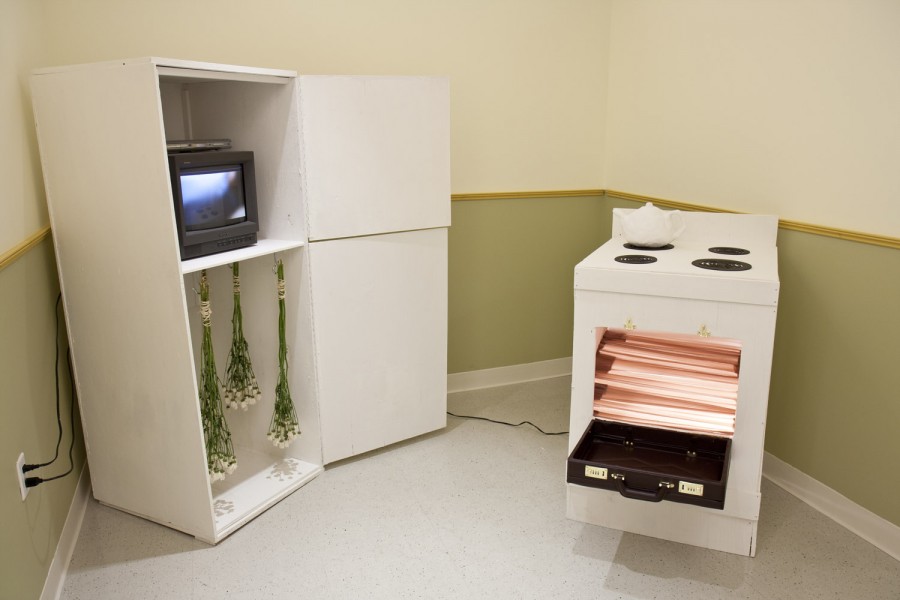 Installation view of a stove with a white tee-pot on it and the oven door open, a cabinet with two storage parts door open and on the top part is a TV with an image on it, on the bottom part are a few flower bouquet hung upside down