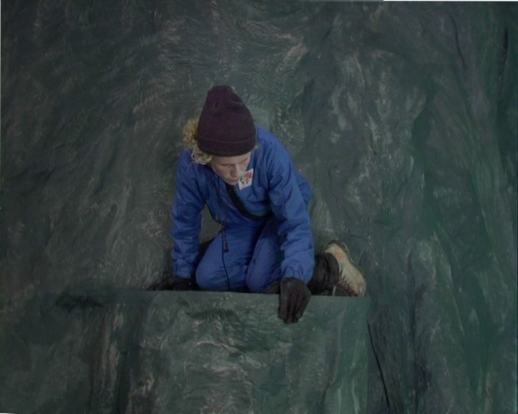 A person with a blue suit, a brown hat, and gloves is sitting on the edge of a cliff, looking down