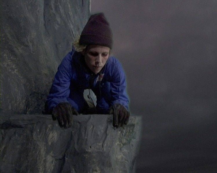 A person with a blue suit and brown hat, and gloves is looking down over a cliff edge
