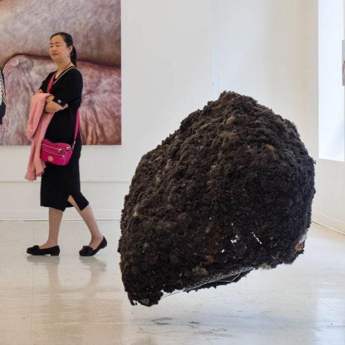 Installation view of a large scale sculpture made of black organic material like human hair, in a room with two people looking around and on the wall in the back is installed a  painting with human body parts