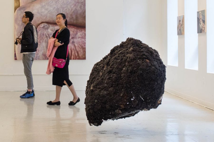 Installation view of a large scale sculpture made of black organic material like human hair, in a room with two people looking around and on the wall in the back is installed a  painting with human body parts 