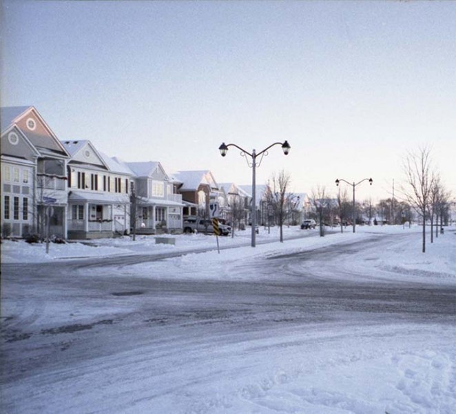 A winter street scene with morning light, houses on the left side, street lights, and a layer of snow covering the scene