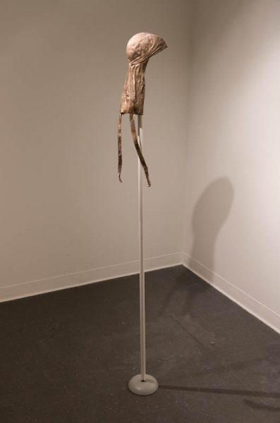 An organic brown shaped sculpture is installed in the corner of a room on a medium-tall white metal rod