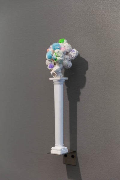A small and thin white column is installed on a tiny shelf, and the column has a bubbly organic shape on top with red, blue, white, green, and other colors.