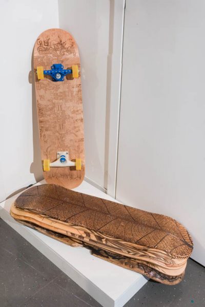 A skateboard is placed in the corner of the room on a white piece of organic material, and near it is a stack of four other wood replacement boards 