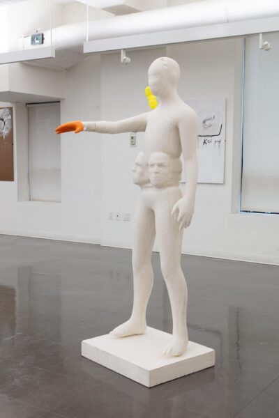 Full-size foam scultpure of a figure with multiple faces around the stomach and a pile of little yellow faces on the right shoulder. Her right arm is extended and the hand is painted in bright orange.