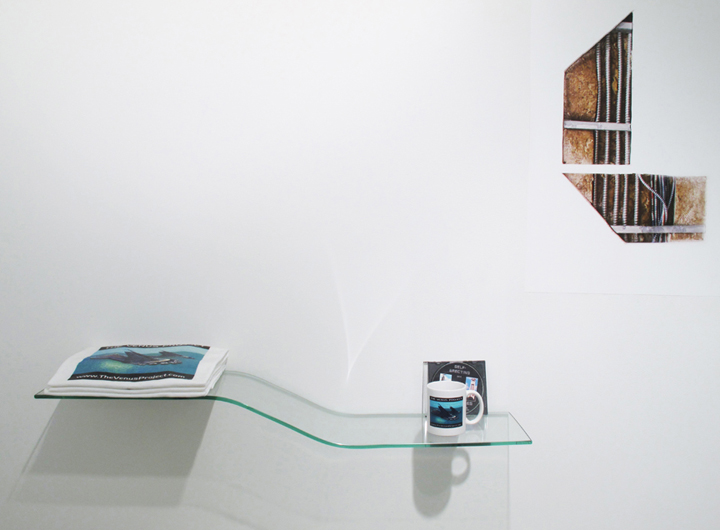 A glass shelf mounted on the white wall with a mug and an illustration behind it and a stack of magazines. On the right side of the shelf is a print illustrating a miscellaneous object 