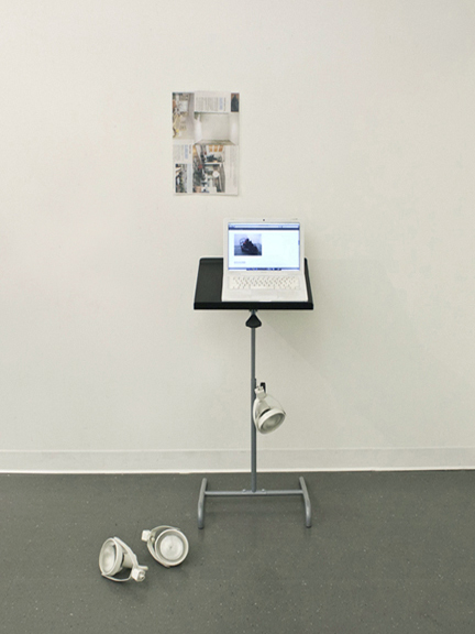 Installation of a white laptop with an image displayed sitting on a stand, a poster on the wall above the laptop, and two white objects placed on the floor in front of the stand