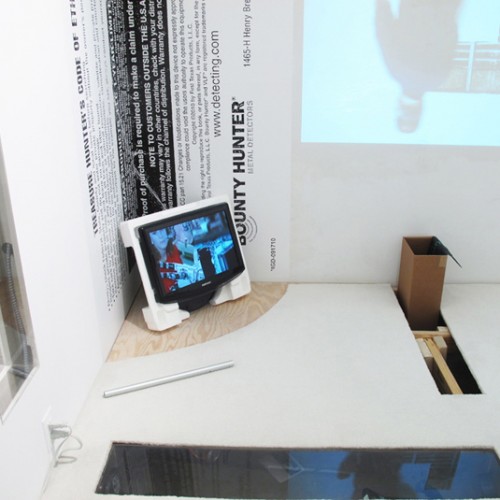 Installation of mixed media including an image projected on the wall, a TV screen with a picture on it, a cardboard box, and in the corner is a vertical sticker applied to the wall with text on it