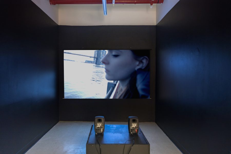 Installation view of artwork by Alexandra Russo. View of a video projection on a black wall of a closeup of a womans face with two audio speakers in the foreground.