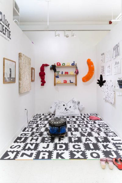 Installation view of miscellaneous sculptures made of fabrics and other organic materials installed on the walls, and the floor is covered with foam with letters, in black and white