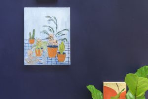 Detail shot of a studio with walls painted deep blue and a painting of a hand above a painting of cacti above a plant.