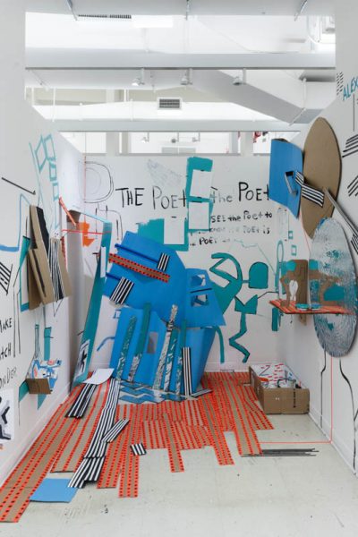 Installation view of blue cardboard sculpture with angles and cutouts, orange tapes on the floor, blue and cardboards on the wall, and other miscellaneous shapes