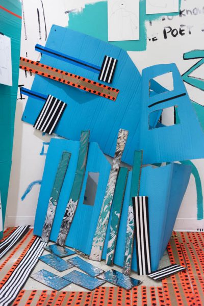Installation view of a blue sculpture made of cardboard with different painted stripes coming from the bottom and an angle in turquoise. the cardboard has cutouts and is cut with angles on the exterior parts