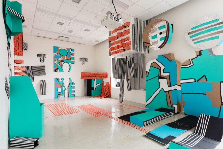 Installation view of an artwork made of cardboard painted on turquoise, blue, and white with some black outlines, installed on the floor and the walls. It is composed of different shapes cut from cardboard, rectangular, rounded line shapes, circles, etc. Ther are orange tapes stick to the floor and on the wall in rectangular shapes, and horizontal lines