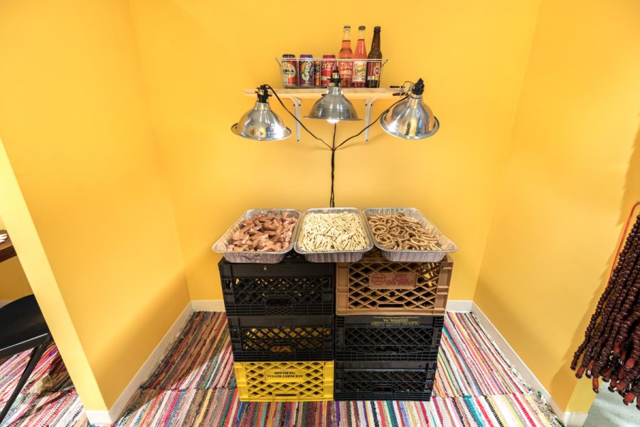 Installation view of artwork by Alize Santana. Three aluminum food trays filled with ceramic fabricated food with lights mounted above sitting on plastic milk crates. Jars of hot sauce a wall mounted rack.