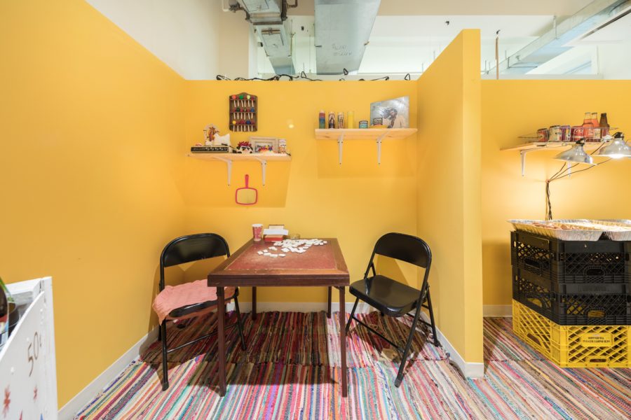 Installation view of artwork by Alize Santana. A breakfast nook created in a space. A table with chairs, shelves on a yellow wall with items on them.