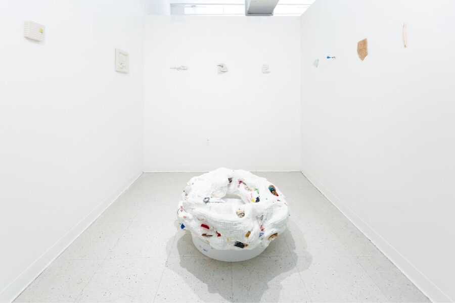 A large white organic torus sculpture installed on the floor made out of small tiles with attached colorful elements