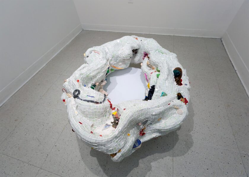 A large white organic torus sculpture installed on the floor made out of small tiles with attached colorful elements