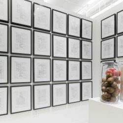 Installation view of a grid with project papers installed, each in its own black frame, and a pedestal with three jars, one with kiwi, one with tomatoes, and one with cereals