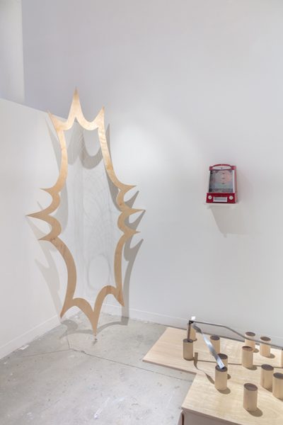 Installation of artwork by Andrew Lee. Geometric wooden shape in the corner of a space, an etch a sketch mounted to the wall, and cylindrical objects on a wooden surface.