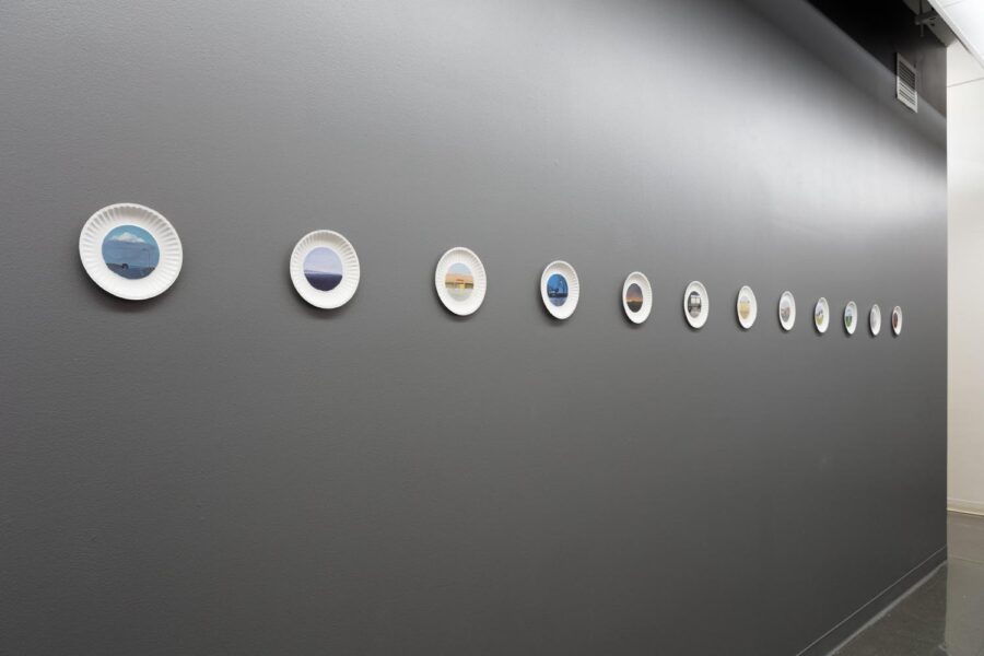 A series of 12 paintings on paper plates installed on a row in a grey wall.
