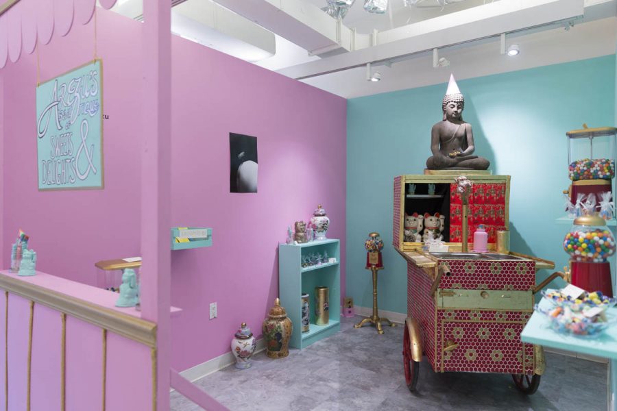 Installation view of a room with candy machines, small statues, ceramic vases and recipients, a pink wall, and a turquoise one. 