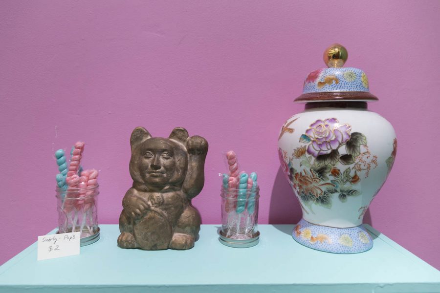 Installation of two jars with Swirly Pops with the price-tag $2, a small brown statue in the shape of a bear with a human face, and a bigger vase with a lid which is decorated with purple flowers, green leaves, and gold outlines for each shape. They are placed on a turquoise surface and the wall behind is pink