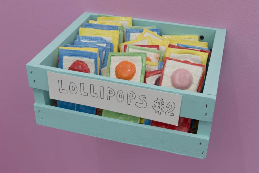 Ceramic sculptures of lollipops painted with vivid orange, red, pink for the candy, and the painted wrap is with blue, green, and red outlines. Al sculptures are in a small turquoise wood crate installed on a pink wall as a shelf. The price tag for candies says Lollipops $2