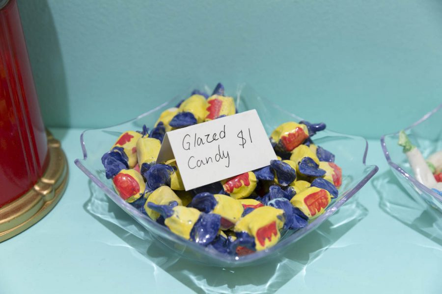 Ceramic sculptures of candies are painted like they are wrapped in yellow with blue and red foil. The candies are in a transparent glass bowl with a name tag made of paper with the text Glazed Candy $1. The table and the wall behind the bowl is turquoise
