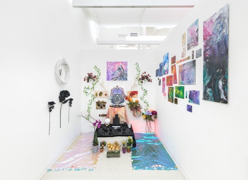 View of an installation by Artemis Razzberry. Multiple abstract multicolored paintings mounted on white walls, various plant foliage and an alter with different items placed around and on top. 