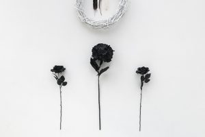 Detail image of an installation by Artemis Razzberry. Multiple black flowers below a white wreath with two feather placed inside.