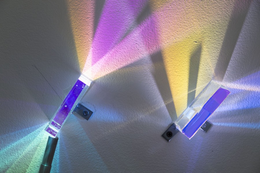 Two glass prisms installed on the wall. Each of them has a light near it and illuminating trough, casting colored light on the wall