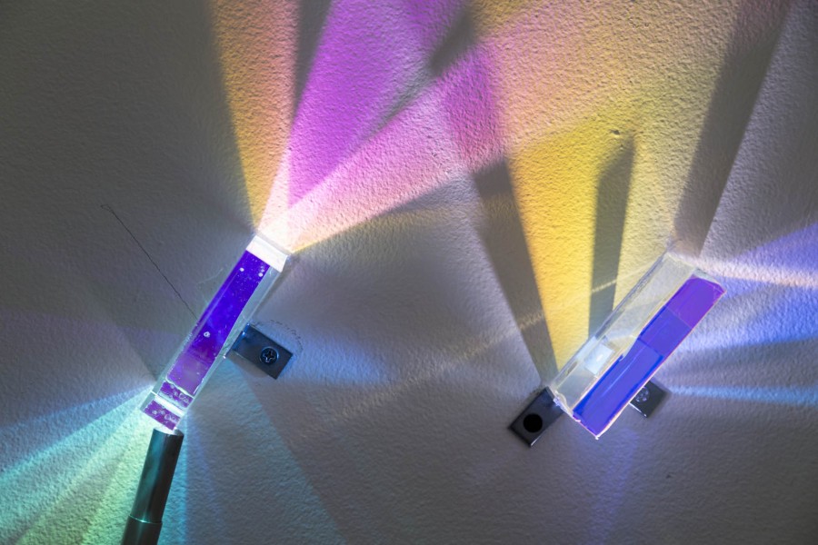Two prisms are installed on a wall with a light source at one end of each prism, making them cast rainbow-like colors on the wall, like yellow, pink, violet, green, etc.