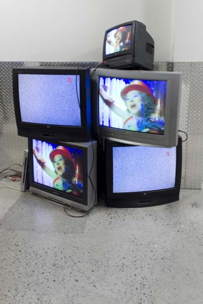 An arrangement of TVs in different shapes and colors in the corner of a room with images of a person with a red hat and two TV's have static images on the screen