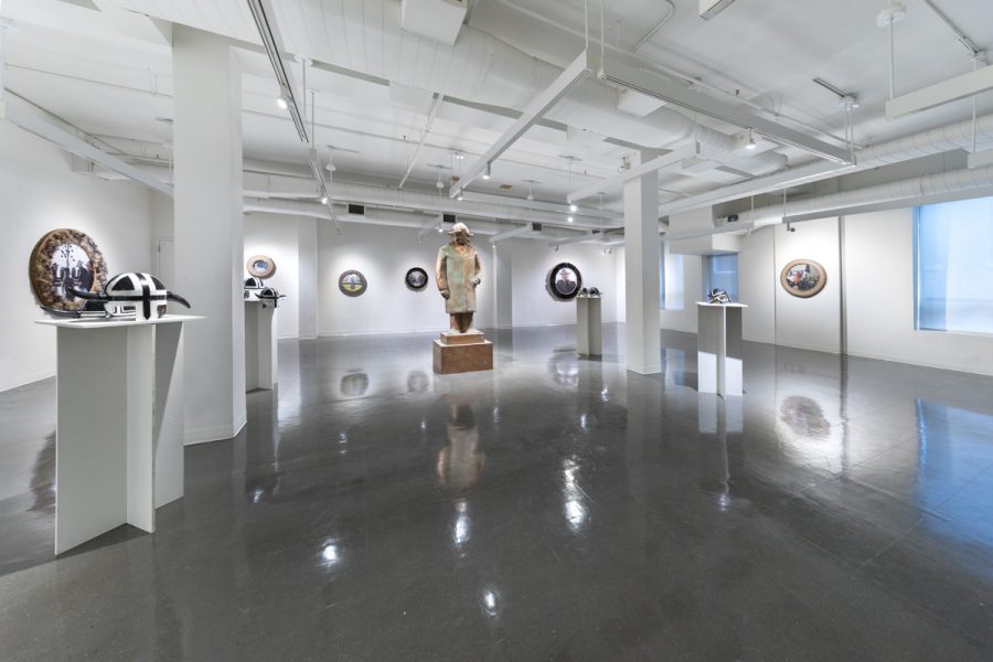 Installation view of artwork by Bjorgvin Jonsson. Multiple circular constructed frames with images of people wearing helmets. Two black and white helmets placed on a pedestal. Large sculpture of a figure wearing a helmet.