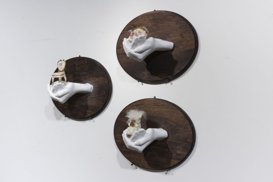 Three white sculptures installed on the wall with a dark wood circle piece, each of two hands holding miscellaneous objects