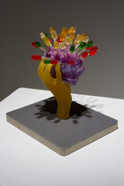 An orange hand sculpture golding up a violet rounded object with jelly teddy bears inserted in it with scouts. the hand is installed on rectangular grey plywood on a white table