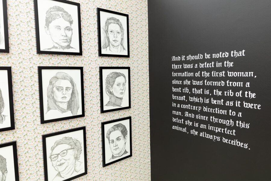 Installation view of a corner with a grid of pencil framed portraits and white paragraph on a black wall that reads: "And it should be noted that there was a defect in the formation of the first woman, since the was formed from a bent rib, that is, the rib of the breast, which is bent as it were in a contrary direction to a man. And since through this defect she is an imperfect animal, she always deceives"