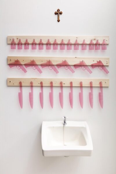 Installation view with a religious cross, rows of poison bottles, guns and large kitchen knifes made out of pink lasetcut plexiglass, and a white sink.