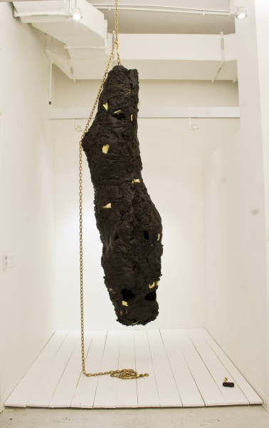 Organically shaped sculpture made with black material hung on the ceiling with a rope and a wooden board white floor.