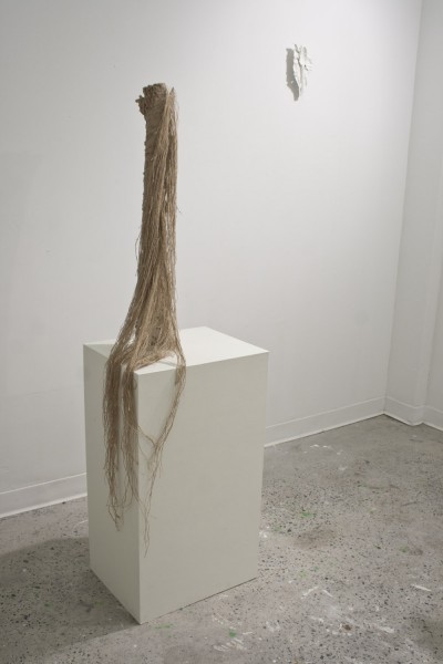 A statue made with long brown organic material on a thin, rounded staple mounted on a big white stand and a small white sculpture mounted on the back wall.