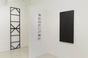 Overview of the installation of artworks by Cameron Richie. This includes a metallic structure on the left side, a series of geometric paintings stacked on the well in the center of the image, and a black painting with geometric shapes.